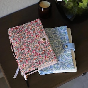 Può includere: Due diari a fantasia floreale con chiusura a bottone. Uno presenta un design floreale rosa, rosso e arancione, mentre l'altro ha un motivo floreale blu e verde. Entrambi sono su una superficie marrone scuro.