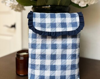 Può includere: Una custodia per libri a fantasia gingham blu e bianca con bordo a balze blu navy. La custodia è appoggiata su una superficie marrone, con un vaso di fiori bianchi sullo sfondo.