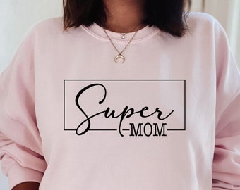 May include: A light pink sweatshirt with the words "Super Mom" in a black, cursive font inside a black rectangle. The sweatshirt is a solid color and has long sleeves. The text "BeChic SVG Graphic Designs" is in the bottom left corner.