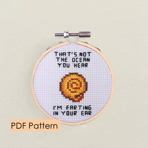 Op de afbeelding: Een witte borduurring met een kruissteekontwerp met een schelp en de tekst "That's not the ocean you hear I'm farting in your ear".