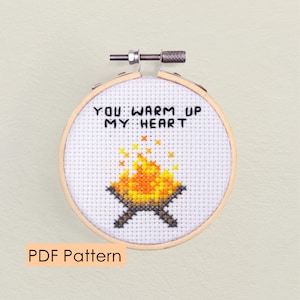Könnte beinhalten: Ein weißer Kreuzstich-Reifen mit einem Lagerfeuer-Motiv und dem Text "You warm up my heart". Der Reifen ist aus Holz und hat einen Metallring.