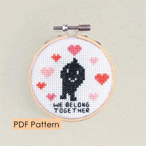Puede incluir: Un patrón de punto de cruz con una silueta negra de un monstruo con una cara sonriente rodeada de corazones rosas. El texto "We Belong Together" está bordado debajo del monstruo.