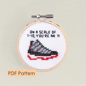 May include: A white embroidery hoop with a cross-stitch design of a black and grey sneaker with red accents. The text "On a scale of 1-10, you're an 11" is stitched above the sneaker.