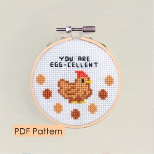 Op de afbeelding: Een witte borduurring met een kruissteekontwerp van een bruin kippetje met een rode kam en de tekst "You are egg-cellent".