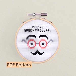 May include: A white cross-stitch embroidery hoop with a black and red design of a face with glasses and a mustache. The text "YOU'RE SPEC-TACULAR!" is stitched above the face.