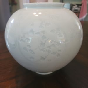May include: A round, off-white ceramic vase with a wide opening. The vase features a detailed, low-relief design of birds and foliage. The design is centered on the vase and is surrounded by additional etched patterns. The vase sits on a dark wooden surface.
