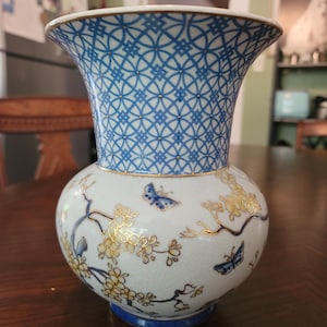 Könnte beinhalten: Dekorative Keramikvase mit weißem Korpus, blauem geometrischen Muster am Hals und goldenen Blumen- und Schmetterlingsakzenten. Die Vase hat einen ausgestellten oberen Rand und einen runden Sockel, geeignet für die Inneneinrichtung.