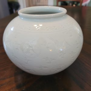 May include: A round, off-white ceramic vase with a wide opening. The vase features embossed decorative patterns and images of birds and flowers. The vase sits on a dark wooden surface.