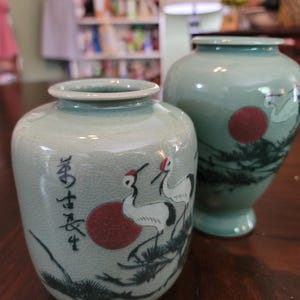 Könnte beinhalten: Zwei Vasen aus Keramik in Celadon-Farbe. Die kleinere Vase zeigt eine rote Sonne, Kraniche und schwarze Kiefernzweige. Die größere Vase hat ein ähnliches Design. Beide Vasen haben eine Craquelé-Glasur.
