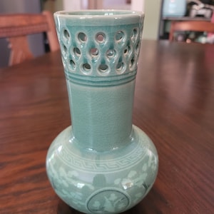 Puede incluir: Un jarrón de cerámica de color celadón con base bulbosa y cuello cilíndrico. El cuello presenta un diseño calado con numerosos agujeros pequeños. La base está decorada con un patrón floral y un emblema circular. El jarrón tiene un esmalte craquelado.