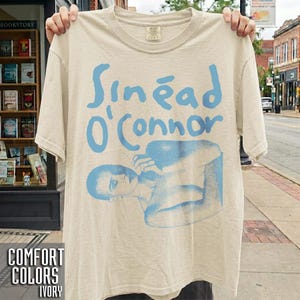 以下が含まれることがあります： アイボリーの半袖Tシャツ。胸元には「Sinéad O'Connor」の文字と、腕を前で組んだ坊主頭の人物のイラストがライトブルーのグラフィックプリントで施されています。