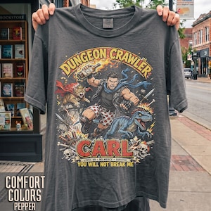 May include: A charcoal grey t-shirt featuring a graphic print of a character labelled CARL, a cat in a cape, and a blue dinosaur. The text reads DUNGEON CRAWLER, CARL, LEVEL 99 NO MERCY SURVIVE, and YOU WILL NOT BREAK ME. The design includes flames and skulls.