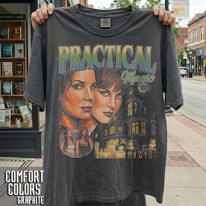 Könnte beinhalten: Ein anthrazitfarbenes T-Shirt mit einem Grafikdruck und dem Text Practical Magic. Das Design zeigt Porträts von zwei Personen, ein großes Haus und eine Gruppe von Menschen, die Kerzen halten. Das T-Shirt hat einen Rundhalsausschnitt und kurze Ärmel.