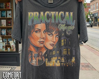 May include: A charcoal gray t-shirt featuring a graphic print with the text Practical Magic. The design includes portraits of two individuals, a large house, and a group of people holding candles. The shirt has a crew neckline and short sleeves.