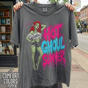 Puede incluir: Una camiseta de manga corta gris carbón con un gráfico de una figura de piel verde y pelo rojo que lleva un top blanco con la palabra GHOUL y pantalones cortos negros. El texto HOT GHOUL SUMMER está impreso en letras grandes rosas y azules.