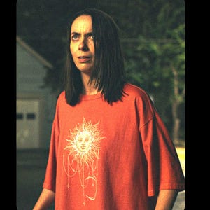 May include: A short-sleeved, crew-neck t-shirt in a vibrant orange hue. The front of the garment features a large, detailed graphic print of a sun with a human face, surrounded by radiating lines, dangling star charms, and a crescent moon shape.