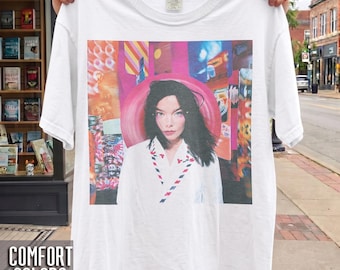 Pode incluir: Uma t-shirt branca de manga curta com uma impressão gráfica de uma pessoa de cabelo escuro a usar uma camisa branca com colarinho e detalhes às riscas vermelhas e azuis, sobre um fundo abstrato vibrante em rosa, laranja e azul.