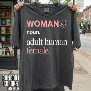 May include: A charcoal gray short-sleeve t-shirt featuring the text "WOMAN noun. adult human female." printed in light pink and white, accompanied by a white speaker icon. The shirt has a crew neckline and a standard fit, made of cotton fabric.