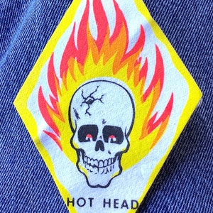 May include: A diamond-shaped patch with a skull design, red eyes, and flames in red, orange, and yellow. The text "HOT HEAD" is below the skull. The patch is set against a blue denim background.