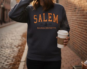 Op de afbeelding: Een antracietgrijze sweater met ronde hals en de oranje tekst SALEM in een collegiaal lettertype, met daaronder de kleinere tekst EST. 1626 en MASSACHUSETTS, samen met een klein zwart silhouet van een heks op een bezemsteel.