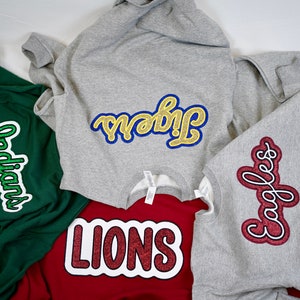 May include: Four sweatshirts with different colored glitter patches. The patches are in the shape of a word bubble and say "Indians", "Tribe", "Lions", and "Eagles".