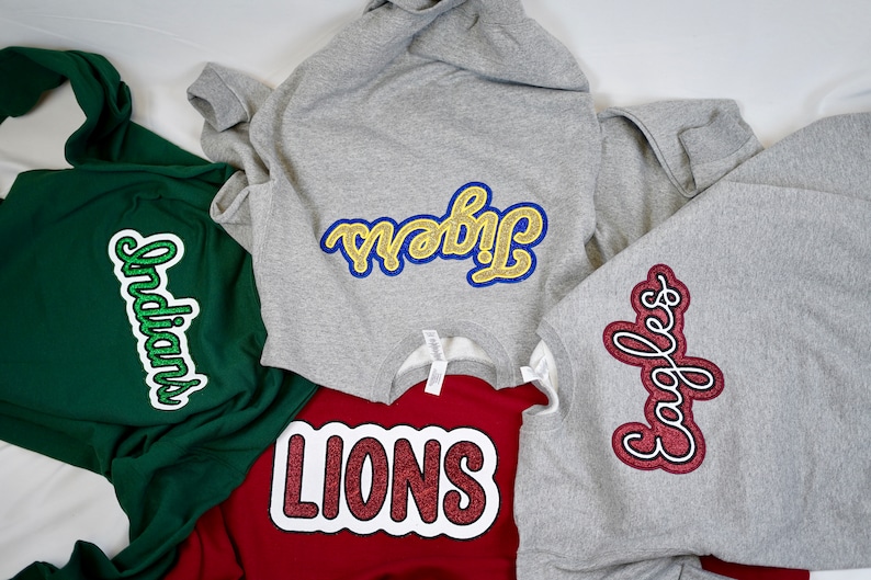 May include: Four sweatshirts with different colored glitter patches. The patches are in the shape of a word bubble and say "Indians", "Tribe", "Lions", and "Eagles".