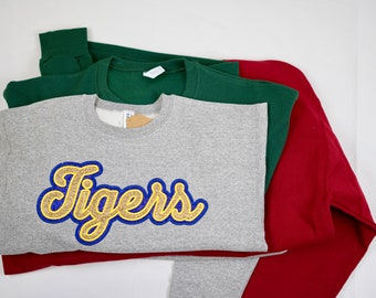 Puede incluir: Tres sudaderas apiladas una encima de la otra. La sudadera de arriba es gris con un gráfico de "Tigers" en dorado y azul. La sudadera del medio es verde. La sudadera de abajo es roja.