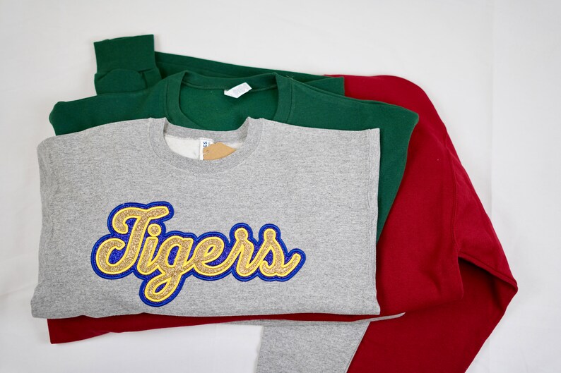 May include: Three sweatshirts stacked on top of each other. The top sweatshirt is gray with a gold and blue "Tigers" graphic. The middle sweatshirt is green. The bottom sweatshirt is red.