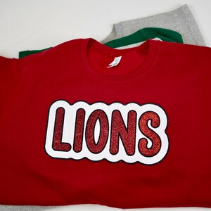 May include: Red sweatshirt with a white outline and red glitter "LIONS" graphic.