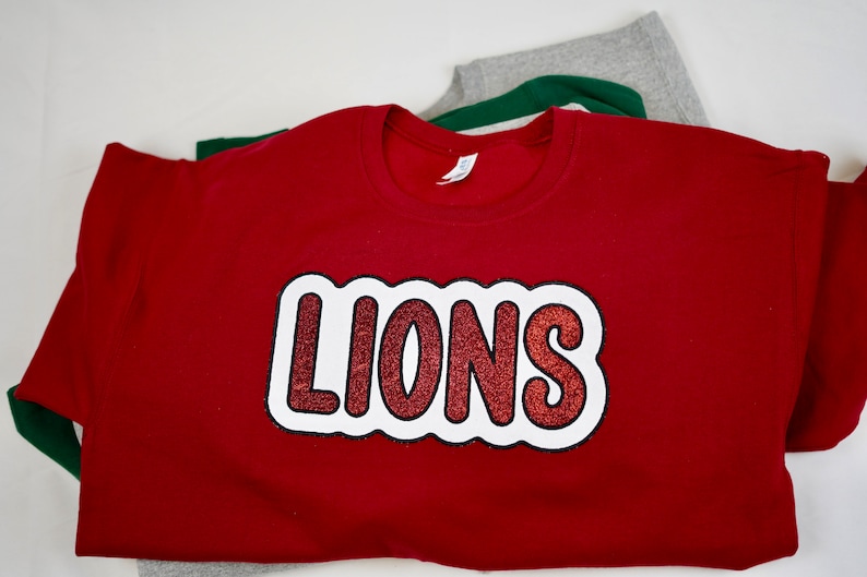 May include: Red sweatshirt with a white outline and red glitter "LIONS" graphic.