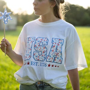 A woman wearing a white t-shirt with the text "USA" and "EST. 1776" printed on it, standing in a grassy field.