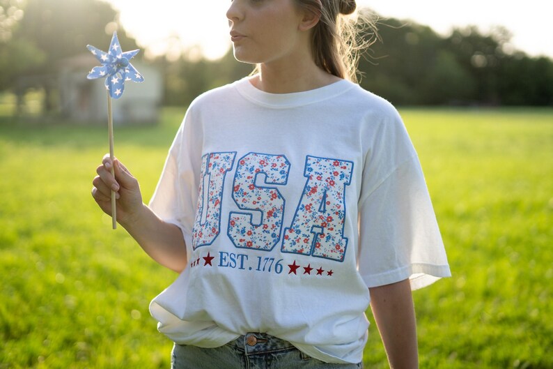 A woman wearing a white t-shirt with the text "USA" and "EST. 1776" printed on it, standing in a grassy field.