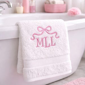 May include: A white cotton bath towel featuring an embroidered design in pink thread. The embroidery consists of a decorative bow at the top and the capital letters MLL centered underneath. The towel has a textured surface and a hemmed border.