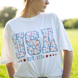 A woman wearing a white t-shirt with the text "USA" and "EST. 1776" printed on it, standing in a grassy field.