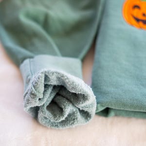 K&ouml;nnte beinhalten: Nahaufnahme der Manschette eines gr&uuml;nen Sweatshirts mit weichem, flauschigem Innenfutter. Der Pullover hat ein K&uuml;rbisdesign auf der Vorderseite.