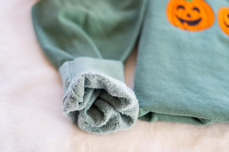 K&ouml;nnte beinhalten: Nahaufnahme der Manschette eines gr&uuml;nen Sweatshirts mit weichem, flauschigem Innenfutter. Der Pullover hat ein K&uuml;rbisdesign auf der Vorderseite.