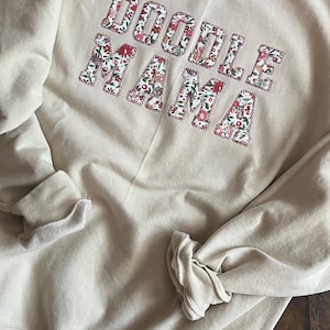 Może przedstawiać: Beżowa bluza z napisem "Doodle Mama" w literach z kwiatowego materiału.