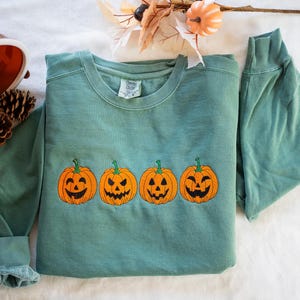 K&ouml;nnte beinhalten: Ein tealgr&uuml;ner Pullover mit einer Reihe von vier gestickten orangefarbenen K&uuml;rbissen mit Jack-o'-Lantern-Gesichtern.