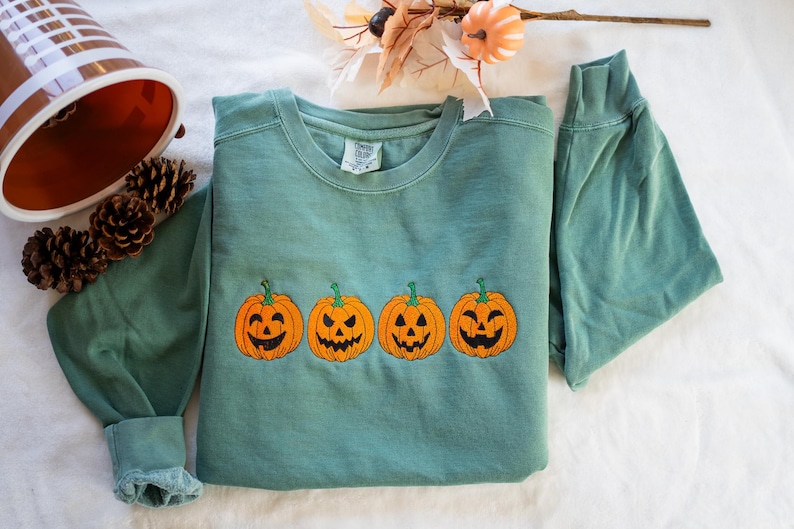 K&ouml;nnte beinhalten: Ein tealgr&uuml;ner Pullover mit einer Reihe von vier gestickten orangefarbenen K&uuml;rbissen mit Jack-o'-Lantern-Gesichtern.