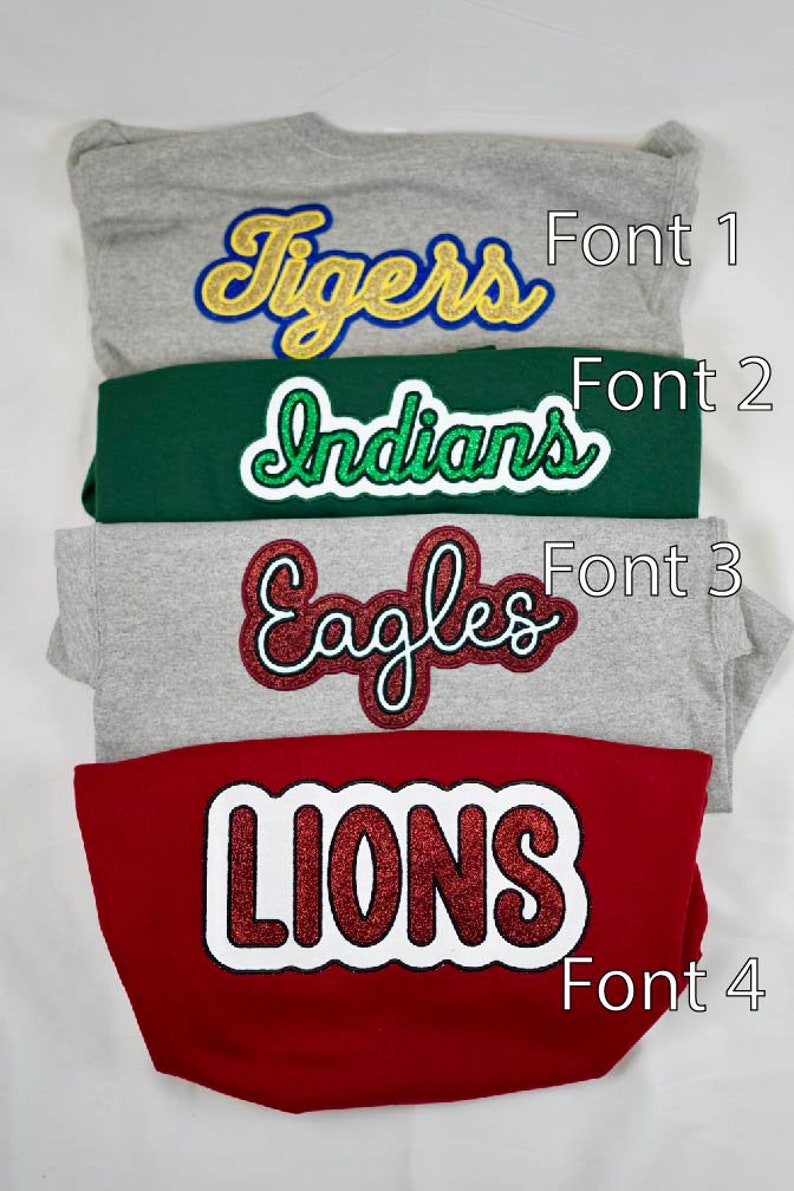 May include: Four sweatshirts with different colored fabric and different glittery iron-on letters. The letters spell out "Tigers", "Indians", "Eagles", and "Lions". Each sweatshirt has a number next to it, indicating the font style of the letters. Font 1, Font 2, Font 3, and Font 4.