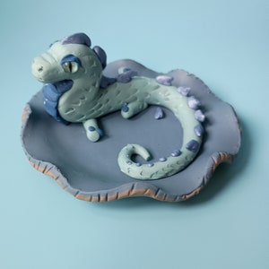 Könnte beinhalten: Eine hellblaue Keramik-Drachenfigur mit violetten Akzenten, die auf einer blauen und braunen Keramikplatte sitzt.