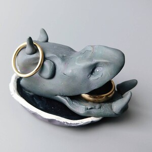 Könnte beinhalten: Ein grauer Keramik-Ringhalter in Form eines Flusspferdes mit offenem Maul. Das Flusspferd hat zwei goldene Ringe in seinem Maul.