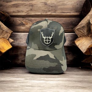 May include: Camouflage trucker hat with a black and white patch featuring a deer head with antlers and a target inside a shield.