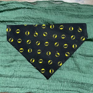 May include: A triangular pet bandana in black fabric featuring a repeating pattern of small yellow Batman logos. The accessory has a straight top edge and a pointed bottom, with the iconic bat symbol printed in yellow across the entire surface.