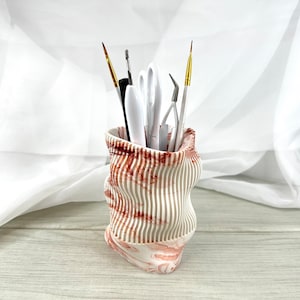 May include: A white ceramic holder with a wavy, textured design and brown marbling. The holder is filled with various tools, including brushes, tweezers, and a nail file.