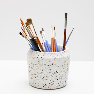 May include: A white ceramic paintbrush holder with a speckled terrazzo pattern. The holder is filled with a variety of paintbrushes in different sizes and colors.