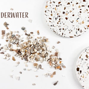 May include: A white background with two round coasters made of white terrazzo with brown and beige flecks. Scattered around the coasters are small pieces of broken seashells in white, brown, and beige. The text "UNDERWATER" is written in brown at the top of the image.