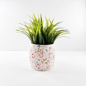 May include: A white ceramic planter with a terrazzo pattern in pink, red, blue, and gold. The planter is filled with green artificial grass.