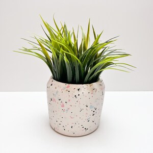May include: A white ceramic planter with a pink, blue, and black terrazzo pattern. The planter is filled with a green artificial grass plant.