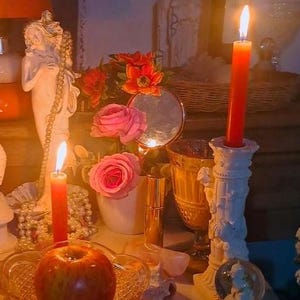 May include: A still-life arrangement featuring lit red candles in ornate holders, a red apple, pink roses, and a small mirror. A white statue and a glass of amber liquid are also present, creating a mystical ambiance.